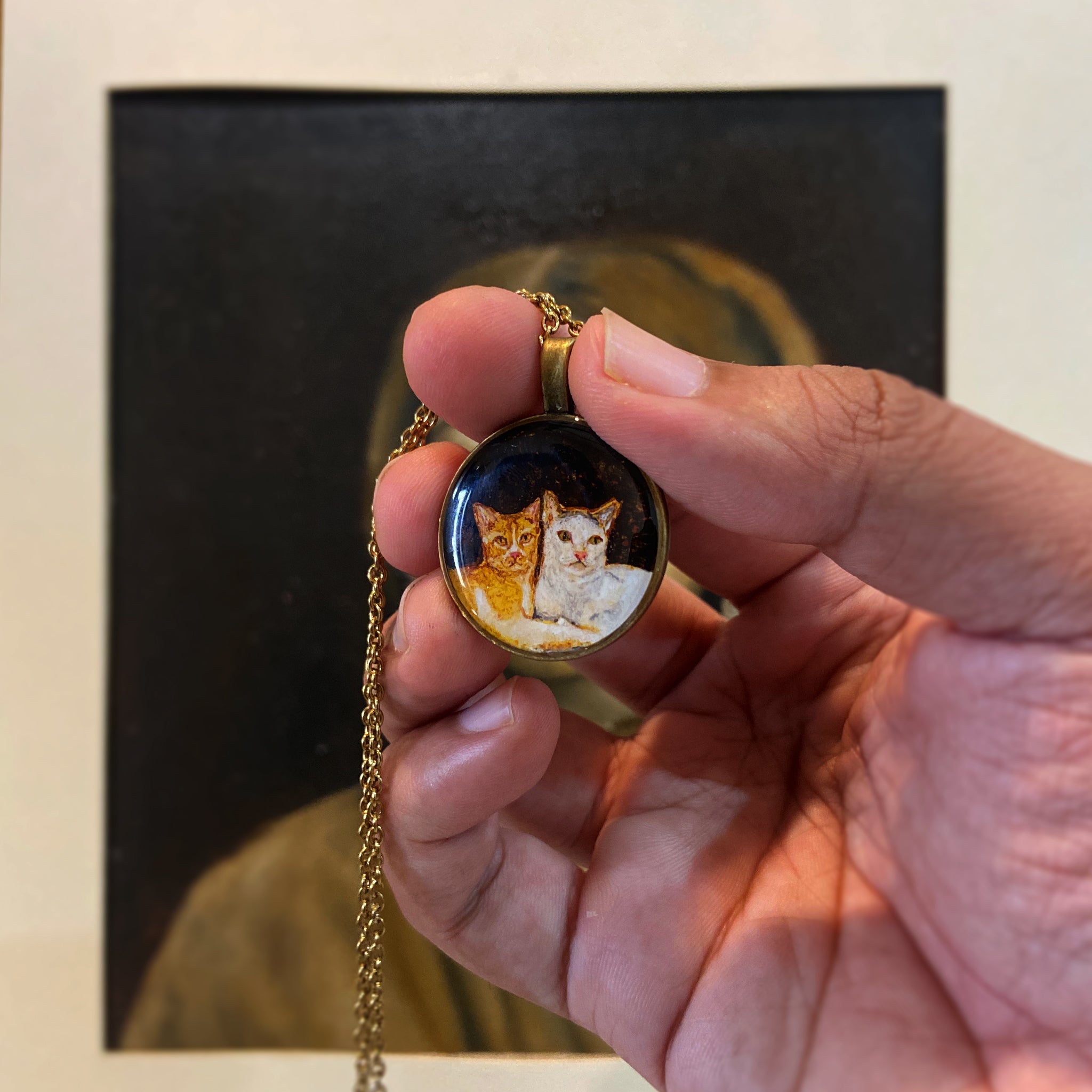 Hand-painted miniature pendant (2.6cm) capturing beloved feline companions: ginger cat alert and attentive with bright amber eyes sitting close to its white companion, who gazes softly ahead with gentle expression, both nestled together against dramatic black background. Vintage-styled portrait held between thumb and fingers with delicate gold chain, coated for dimensional shine, showcasing these cherished pets in intimate detail.