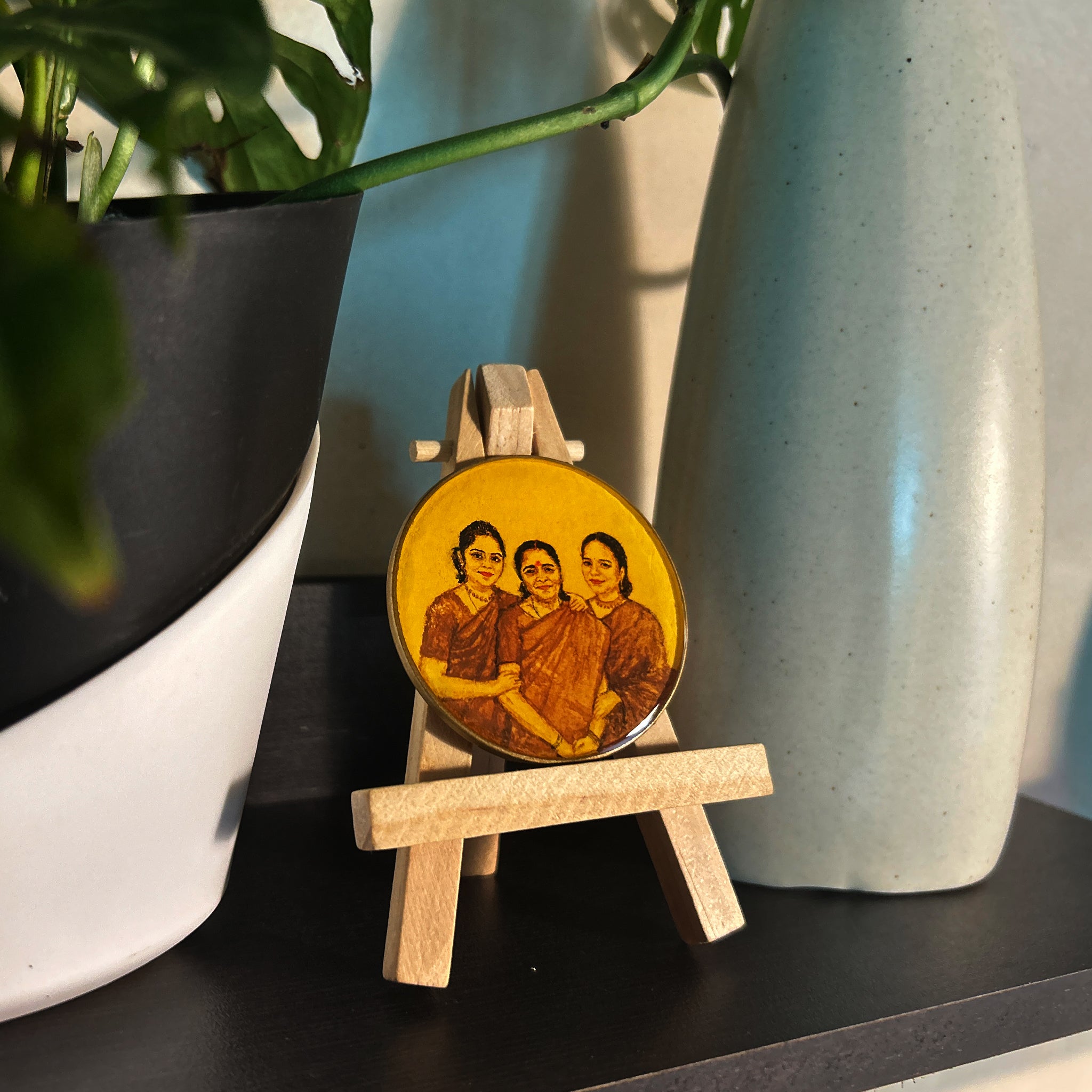 Hand-painted miniature (5cm) portraying loving family trio: mother centered, embracing daughters with tender smile, daughters nestled close displaying warmth in their expressions, all in traditional saris. Rich brown and burnt umber tones on naples yellow background create vintage appeal. Artwork perched on wooden easel amid modern decor - sunlit space with monstera plant casting gentle shadows, demonstrating how this vintage-styled, heirloom-quality piece harmonizes with contemporary home styling.