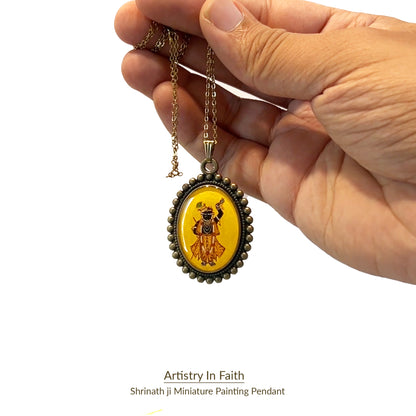 This pendant from the 'Artistry in Faith' collection captures a detailed miniature of Lord Shrinathji, painted in black on a 23mm x 32mm oval base. The deity's white eyes stand out against his black figure, and he wears a yellow dhoti, a golden crown with a green peacock feather, and is adorned with pearls and a garland. The background is a vintage yellow, while the pendant's metal frame features a traditional moti-style border, combining elegance and spiritual significance.