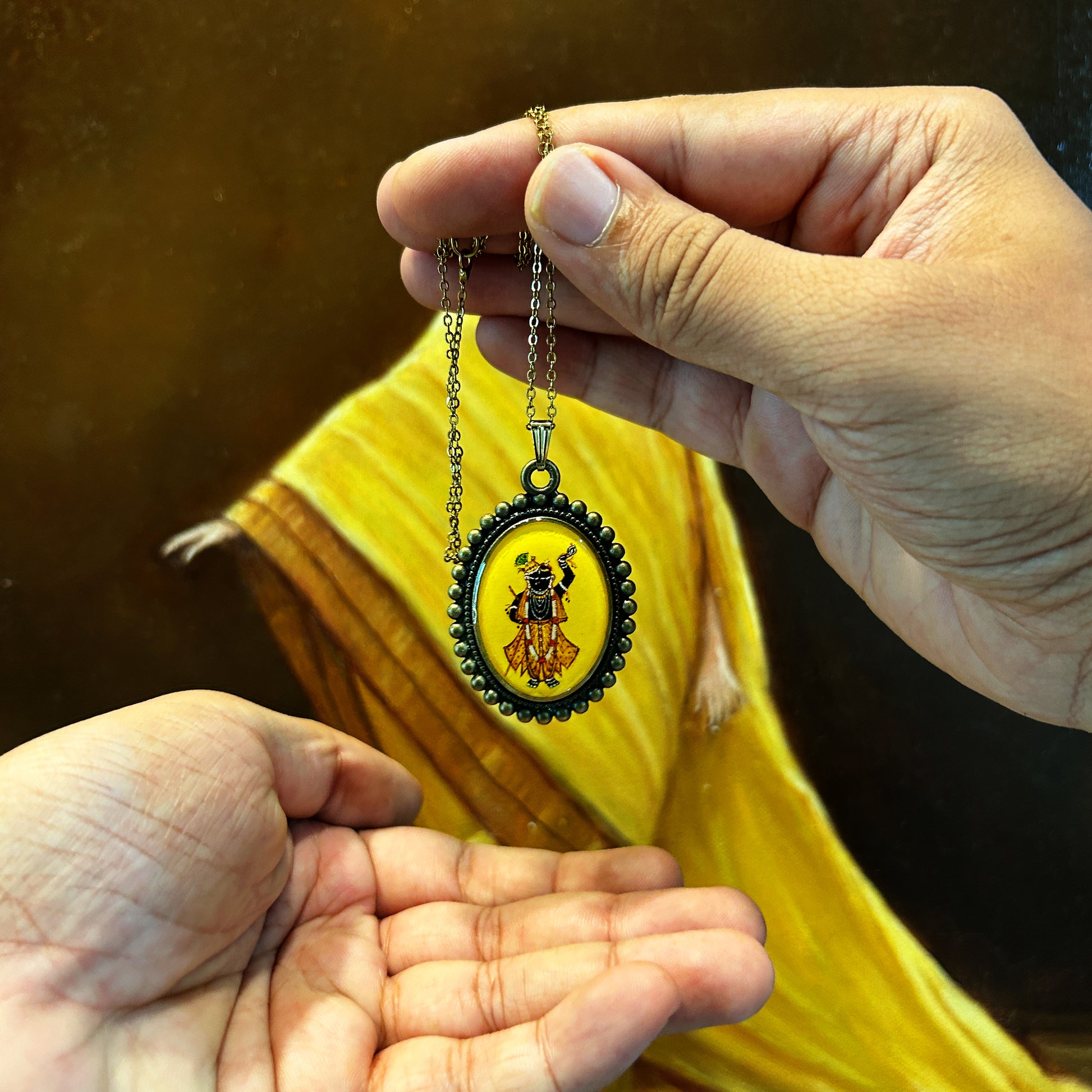 Lord Shrinathji pendant from the 'Artistry in Faith' collection, featuring a 23mm x 32mm oval miniature painting. The deity is depicted in black, with sharp white eyes, wearing a yellow dhoti, one hand raised and the other on his torso. A golden crown with a green peacock feather decorates his head, complemented by a garland and white pearls. The vintage yellow background contrasts with the moti-style metal frame, making this a unique piece of wearable art.