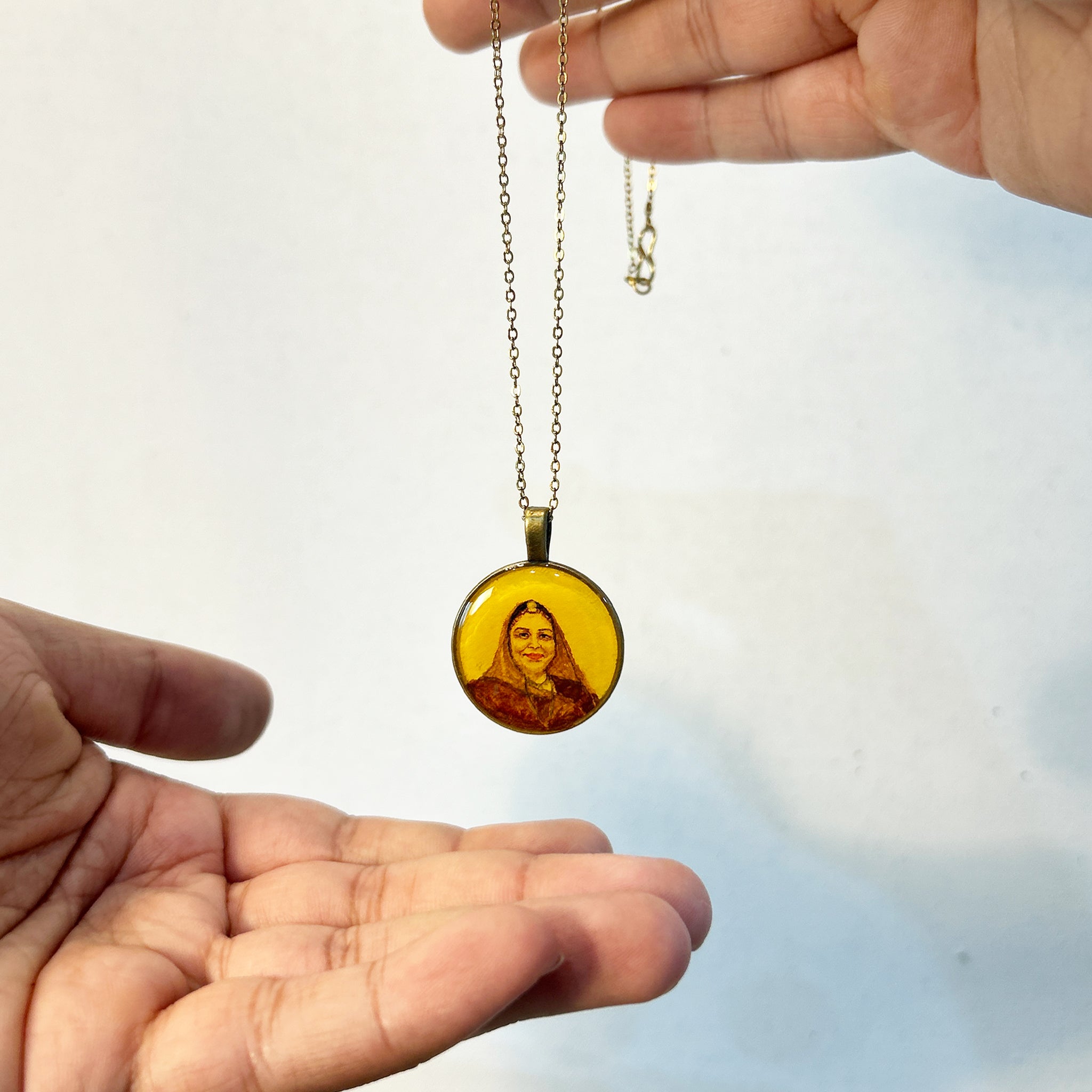 AMAANSA's artisan portrait pendant (26mm), featuring a mother in classic Rajasthani ensemble, highlighting the traditional maang tika and headscarf detail. Her gentle expression rendered in timeless style with vintage-inspired hues. Simple brass border enhancing the cultural elements. Delicately held between fingertips with antique gold colored chain suspended, photographed against soft blue-white background. Sealed with luminous finish, this wearable art piece preserves cultural memories.