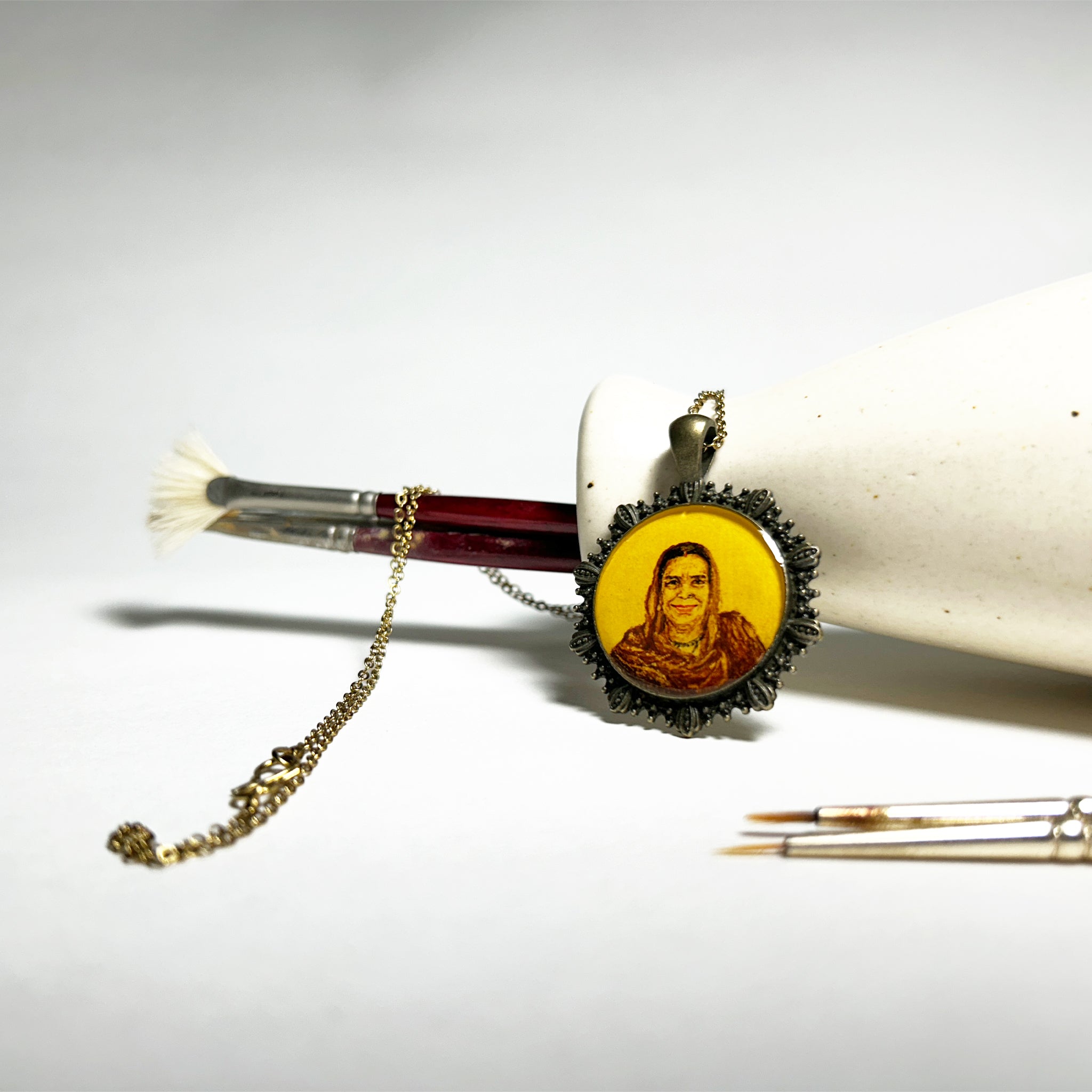 AMAANSA's artisan portrait pendant (26mm), connecting mother and son across oceans. Her warm smile and traditional attire rendered in timeless style with vintage-inspired hues. Embraced by our signature Coastal Shell Frame in antique brass finish. Elegantly suspended beside artist's brushes with antique gold colored chain flowing over brush, set against clean white background. Sealed with luminous protective layer, this wearable treasure keeps loved ones close.