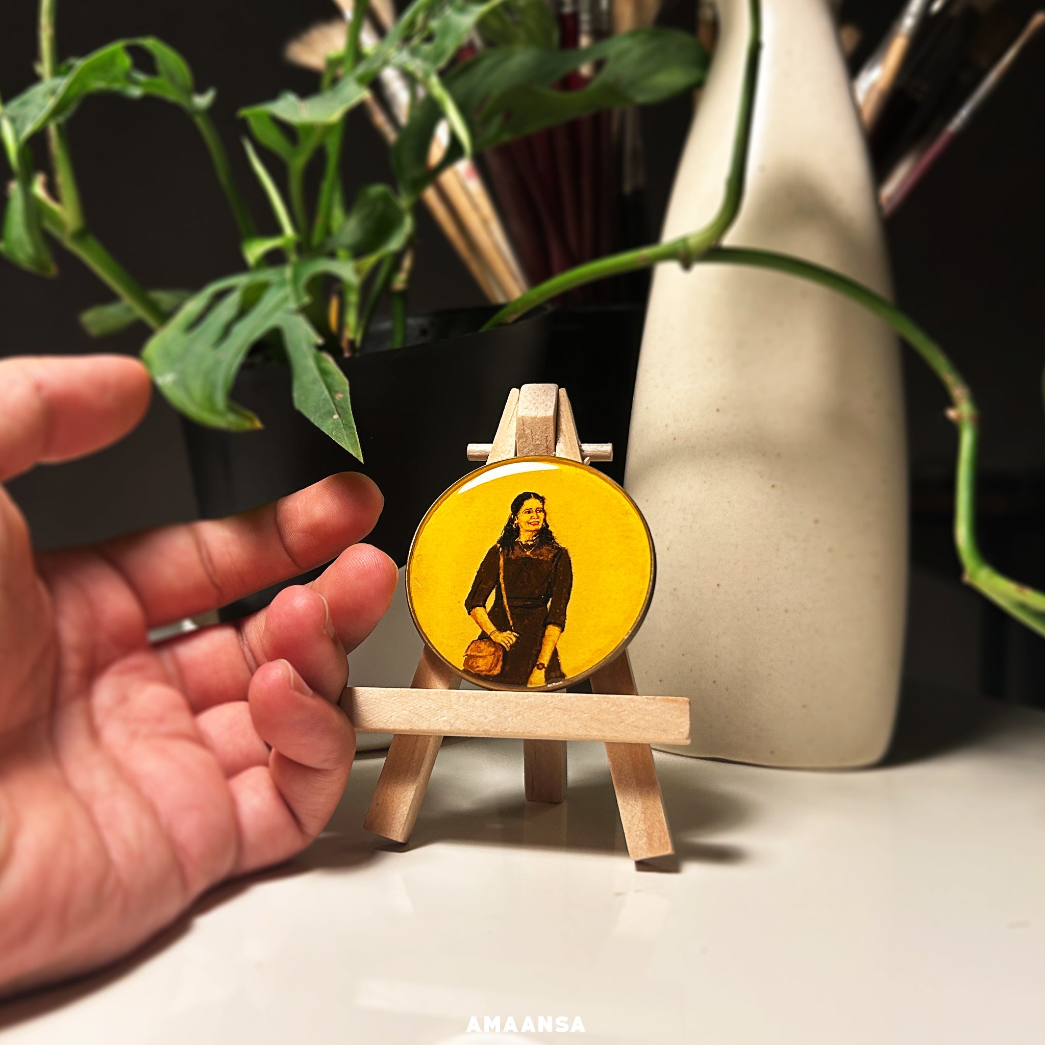 A detailed miniature painting of a woman standing confidently, set against a vibrant yellow background. The portrait, painted on a 5cm diameter circle, shows the woman wearing traditional attire and holding a small bag. The painting is showcased on a small wooden easel, with a hand gently touching the base, emphasizing the artwork's size. In the background, there are green plants and artist brushes, adding a creative studio ambiance. This custom tabletop showpiece from AMAANSA Fine Arts Studio highlights th