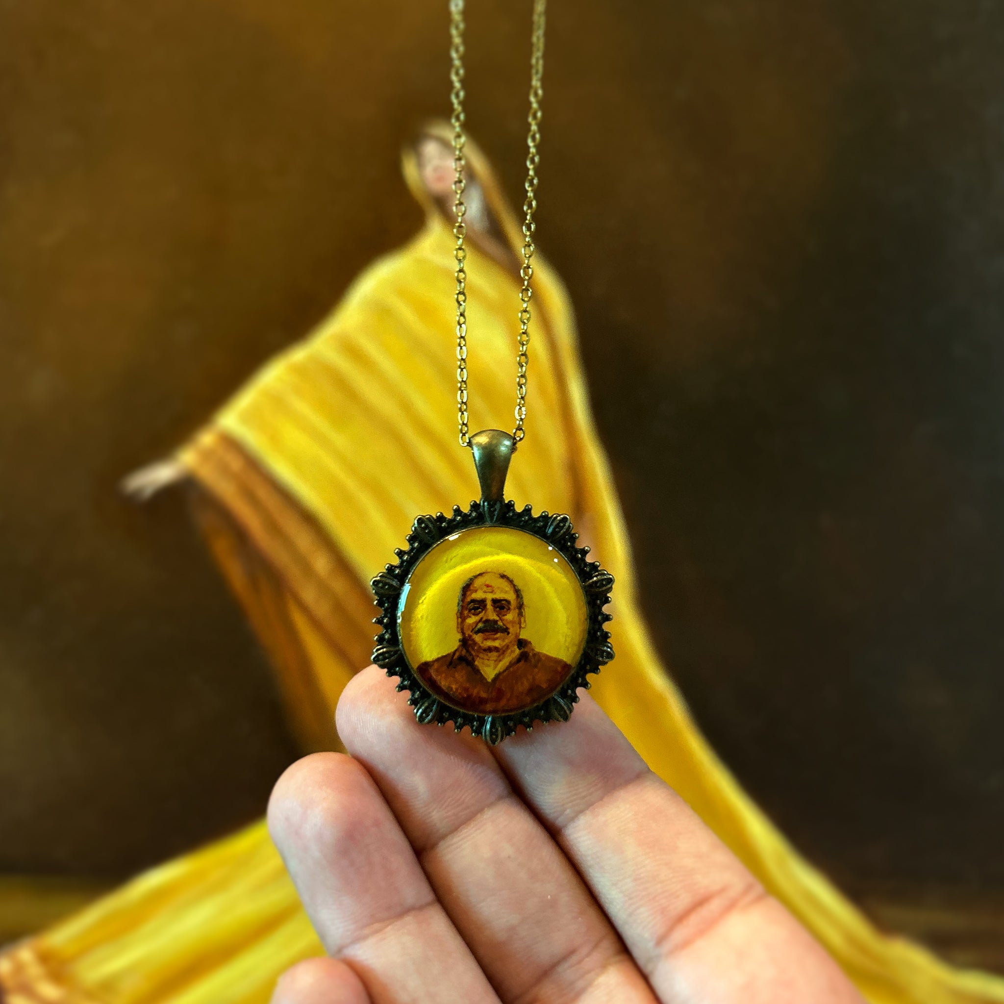 AMAANSA's hand-painted miniature portrait pendant (26mm), preserving a beloved father's warm smile and gentle expression in timeless style. His characteristic features painted with heritage-inspired hues, framed by our signature Ocean Shell Border (named Sea Shells) in antique brass finish. Tenderly held in palm with antique colored chain suspended, set against artist's oil painting featuring draped yellow sari. Protected by crystal-clear sealant, this memorial keepsake keeps a daughter's precious memories 