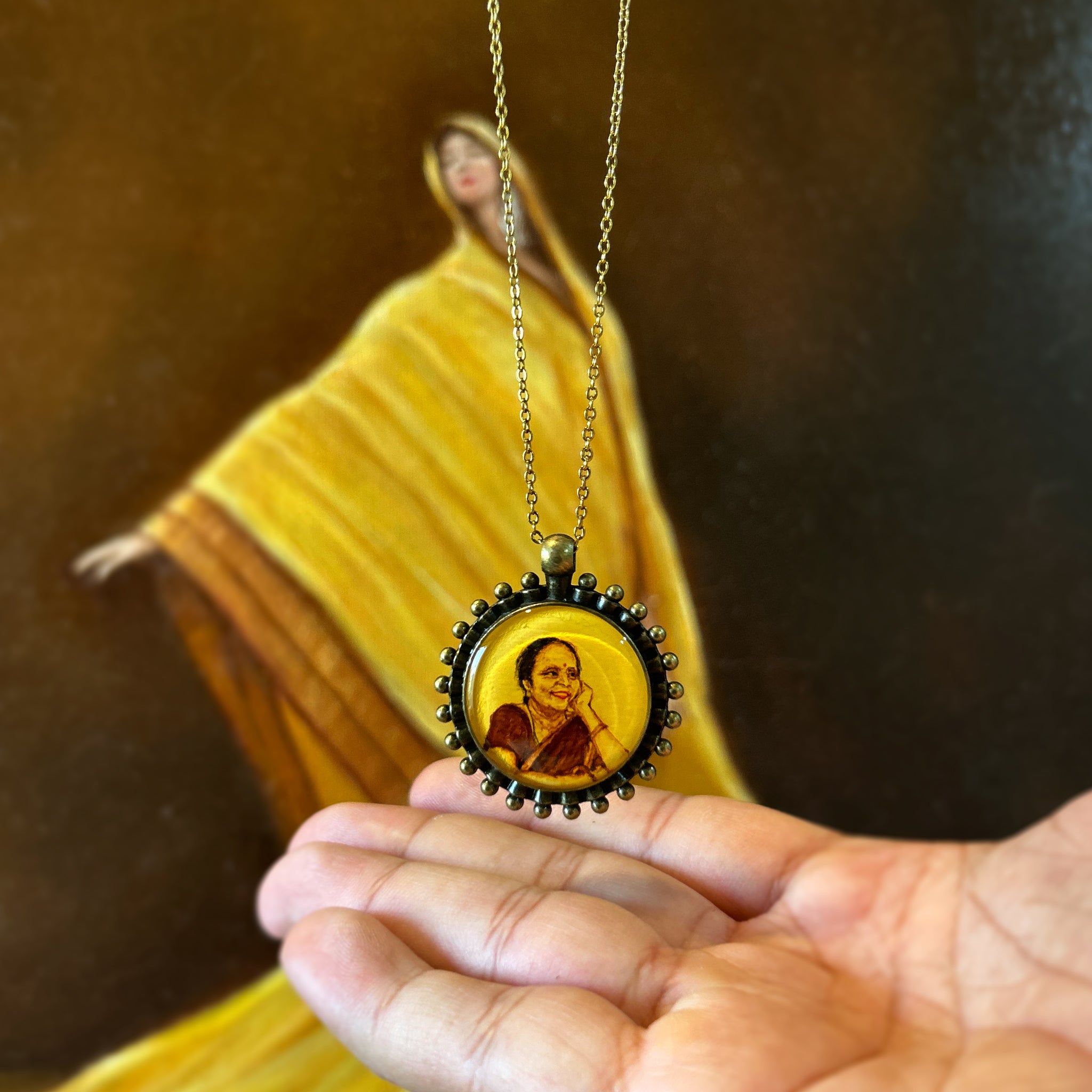AMAANSA's hand-painted miniature portrait pendant (26mm), immortalizing a mother's graceful pose with hand resting on cheek, draped in traditional red saree. Her gentle smile painted with heritage-inspired hues, framed by our signature Heritage Pearl Border (named Pathakkam Moti) antique brass finish. Delicately balanced in palm with antique colored chain flowing, set against artistic yellow silk backdrop. Protected by crystal-clear sealant, this wearable keepsake preserves a daughter's cherished memories.