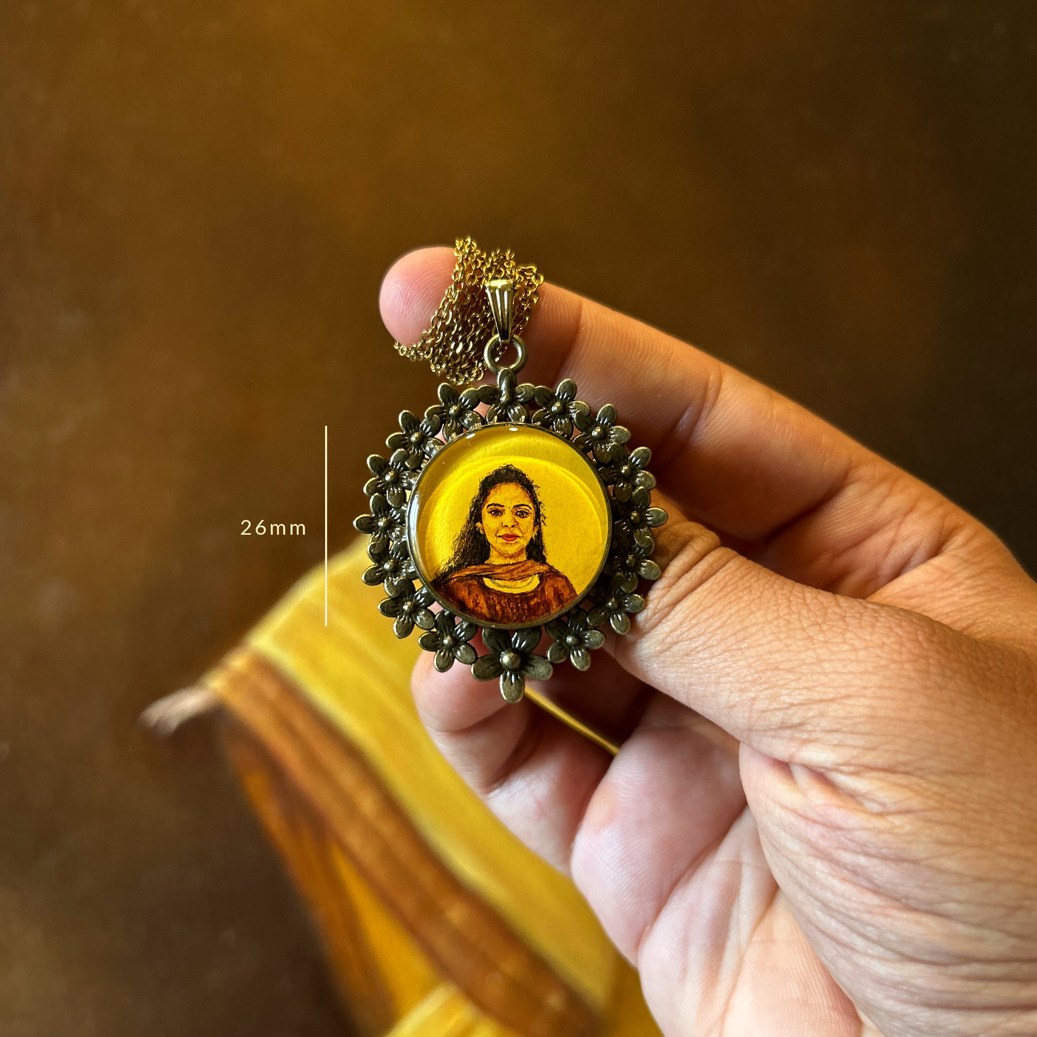 AMAANSA's artisan miniature art pendant (26mm), showcasing a beautiful portrait of a young lady with flowing curls in heritage-inspired hues. Her warm smile painted with artistic precision, framed by our signature Vintage Floral Wreath in antique brass finish. Elegantly presented with antique gold colored chain against draped fabric. Preserved with luminous protective layer, this bespoke jewelry art piece celebrates special moments.