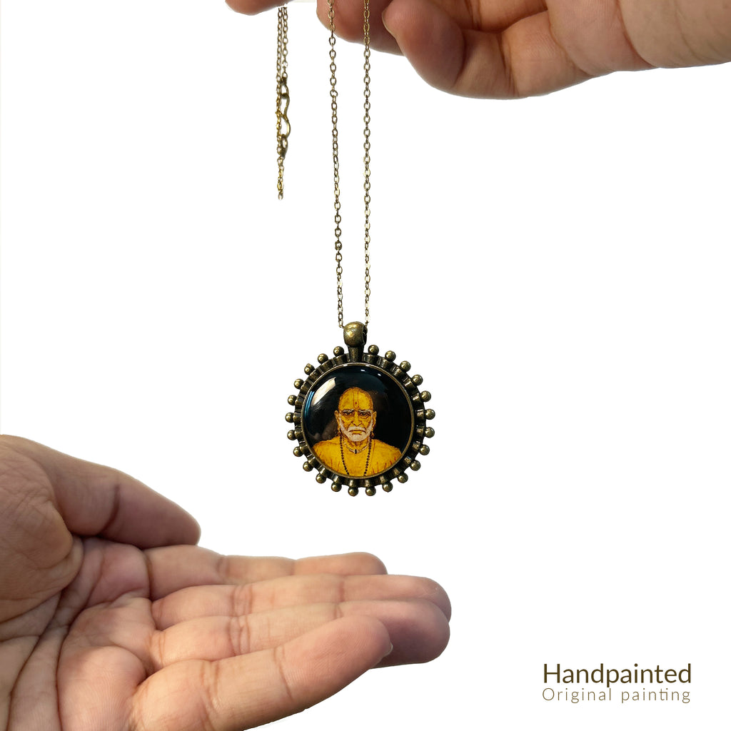 AMAANSA Fine Arts Studio presents: Divine miniature pendant (26mm) from Artistry in Faith, featuring Shri Swami Samarth's sacred portrait. His penetrating gaze rendered with devotional precision, framed by our signature Pearl Border (Pathakkam Moti) in antique brass finish. Pendant gracefully suspended between both palms with antique gold colored chain flowing, captured against pure white backdrop. Protected by crystal-clear sealant, this blessed keepsake serves as a portable shrine.